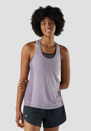 Glimlachende vrouw met krullend haar, gekleed in een lavendelkleurige, mouwloze sporttop en zwarte korte broek, staand met één arm achter haar rug gekruist.