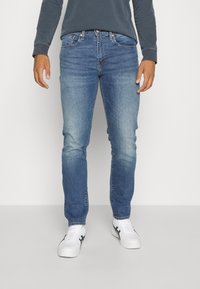 Vaqueros de hombre azul claro hechos de mezclilla, con un corte ajustado, lavado descolorido y diseño de cinco bolsillos. Combinados con zapatillas blancas.