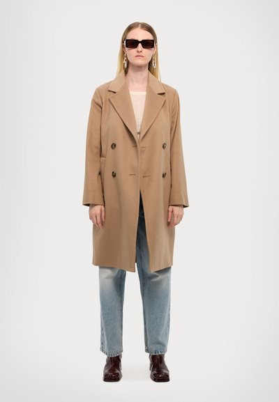 Tan wool blend double-breasted coat with large lapels, dark buttons, side pockets, paired with light blue jeans and burgundy ankle boots.