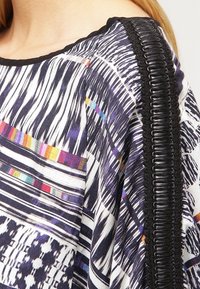 Koszulka tekstylna z wielokolorowymi wzorami geometrycznymi i paskowymi, z czarnym teksturowanym wykończeniem na ramieniu dla dodatkowego akcentu.