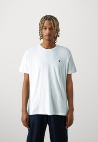 Camiseta blanca de algodón con cuello redondo, mangas cortas y un pequeño logo azul marino en el lado izquierdo del pecho. Combinada con jeans de mezclilla oscura.