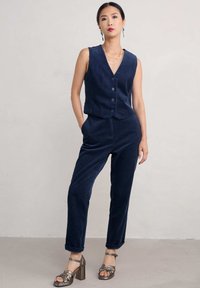 Modra kordna brezrokavnica in hlače s povečanjem v izrez, gumbi z obeh strani spredaj in zavihani hlačnice; v kombinaciji z metaliziranimi petkami.