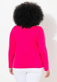 Osoba s kudrnatými černými vlasy má na sobě jasně růžovou dlouhou košili a bílé kalhoty, stojí zády k fotoaparátu na jednobarevném bílém pozadí.