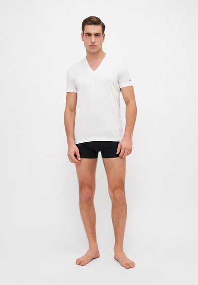 T-shirt blanc à col en V en tissu doux, à manches courtes, associé à un short noir ajusté. Le modèle est pieds nus contre un fond uni.