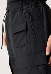 Pantalons cargo noirs en nylon léger. Dotés d'une taille élastique, d'une poche zippée sur le côté et d'une poche à rabat avec fermeture Velcro.