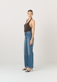 Ciemnobrązowy top na ramiączkach, niebieskie dżinsy z wysokim stanem, szerokie nogawki, jasny odcień denimu, czarne sandały na obcasie, modelka stojąca na jednolitym tle.