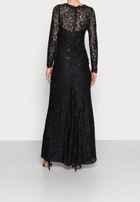 Černá krajková večerní šaty s dlouhými rukávy, květinovým vzorem a flitry. Přiléhavá korzetová část a rozšířená sukně se středovým ventilačním otvorem na zadní straně.