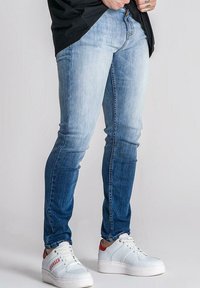 Denim traperice s gradijentnom bojom od svijetloplave do tamnoplave s uskim krojem, koje imaju glatku teksturu i minimalne šavove. Uparene s bijelim tenisicama.