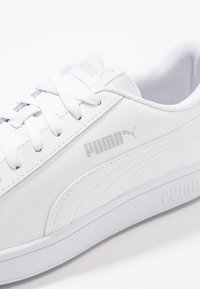 Sneaker sportive bianche realizzate in materiale sintetico. Le caratteristiche includono una superficie texturizzata, lacci bianchi e un logo Puma argentato sul lato.
