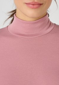 Maglione rosa chiaro a collo alto in texture a costine. Presenta un design aderente con colletto alto e un tessuto morbido, che mette in risalto uno stile minimalista.