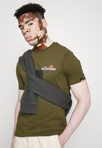 Olivgrünes T-Shirt mit kurzen Ärmeln und einem kleinen gestickten Logo, kombiniert mit einem dunkelgrauen Pullover, der um die Schultern gebunden ist.