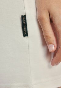 Wit stof met een gladde textuur, met een zwart label waarop in gouden letters "ROCKETT ST. GEORGE" staat, dichtbij een hand.