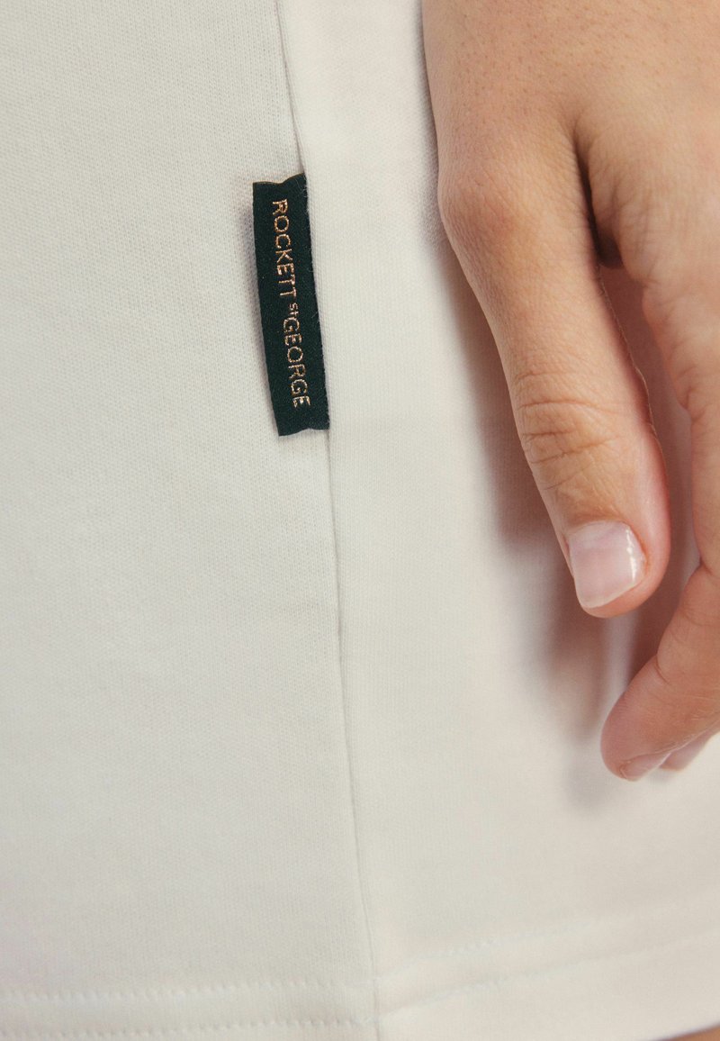 Wit stof met een gladde textuur, met een zwart label waarop in gouden letters "ROCKETT ST. GEORGE" staat, dichtbij een hand.