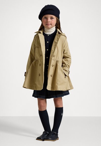 Beige bomullstrenchcoat med en utsvängd design, knäppningar och sidofickor. Matcher med en marinblå tröja, jeaanskjol, knähöga strumpor och skor.