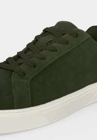 Baskets en daim vert foncé avec lacets assortis et semelle en caoutchouc blanche, vues de côté et légèrement en hauteur.
