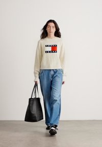 Kremowy sweter z prążkowanej dzianiny z grafiką "TOMMY JEANS", niebieskie luźne dżinsy, czarna torba typu tote i czarne buty z białymi podeszwami.