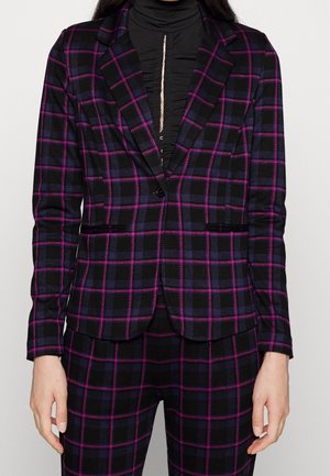 Femme portant un costume noir à carreaux violets et roses, associé à un haut noir froncé à col montant.