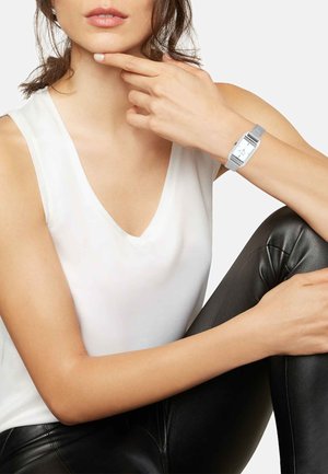 Kvinde iført hvid ærmeløs top og sorte bukser, der rører ved hagen, iført rektangulært sølv mesh ur på venstre håndled.