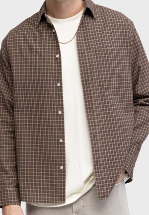 Mężczyzna ubrany w brązową kraciastą koszulę na guziki, założoną na białą koszulkę, noszący złoty łańcuszek i jasne spodnie.