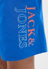 Plave kupaće hlače s glatkom teksturom, sa velikim logotipom "JACK & JONES" u bijeloj i narančastoj boji s lijeve strane.