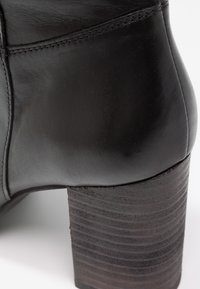 Gros plan sur une bottine en cuir noir avec un talon bloc empilé et des coutures visibles sur la partie supérieure.