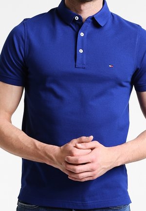 Mand iført en tætsiddende kongeblå kortærmet polo med lille logo på brystet, hænderne samlet ved livet mod en hvid baggrund.
