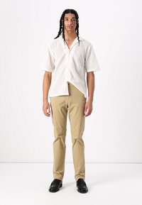 Chemise blanche à manches courtes avec un motif en zigzag, associée à un pantalon slim beige clair et des chaussures noires. Debout sur un fond uni.