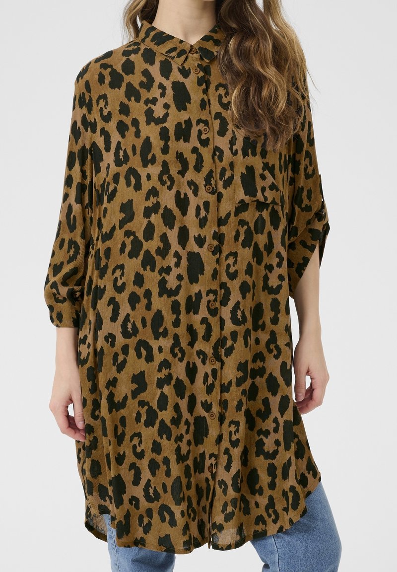 Femme portant une chemise ample marron à imprimé léopard avec boutons et manches trois-quarts, sur un jean bleu, debout devant un fond uni.