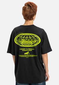 Czarny, oversize t-shirt z dużą fluorescencyjno-żółtą grafiką na plecach, w tym tekstem i wzorem kuli ziemskiej. Krótkie rękawy, okrągły dekolt.