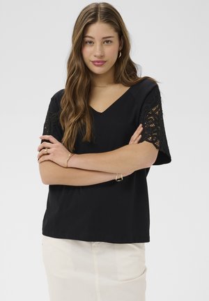 Femme aux longs cheveux bruns portant un haut noir à manches en dentelle et une jupe blanche, les bras croisés, fond neutre.