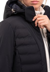Chaqueta negra acolchada con capucha, con cierre de cremallera en el frente y costuras destacadas. El material parece suave con un diseño elegante y moderno.