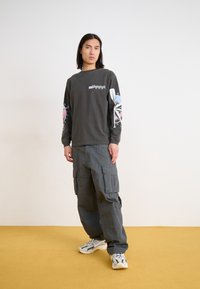 Young person with long hair stands on yellow floor, wearing dark graphic long-sleeve shirt, gray cargo pants, and white sneakers.