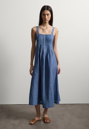 Mujer con vestido midi azul sin mangas con pliegues en el corpiño y sandalias planas marrones, de pie contra una pared blanca lisa.