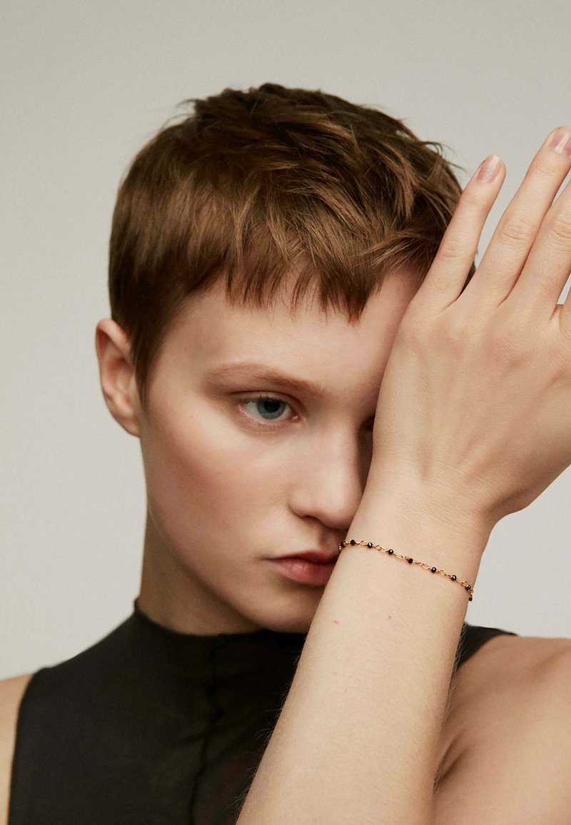 Pulsera dorada con pequeñas cuentas negras, llevada en una muñeca de tono de piel claro. La mano del modelo está levantada mientras viste una blusa negra.
