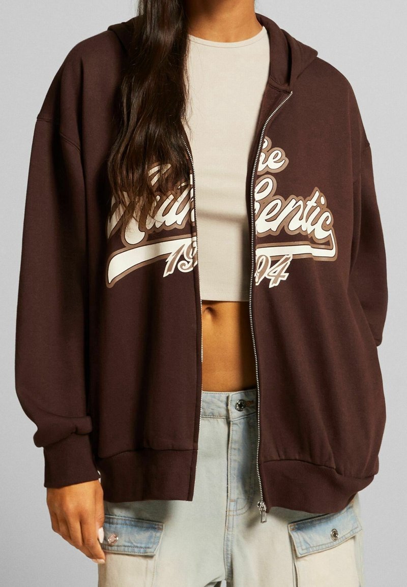 Hnědá mikina na zip s kapucí, s bílým nápisem "Authentic", volný střih a vroubkované manžety, v kombinaci se krémovým topem.