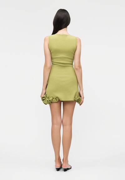 Γυναίκα με μακριά σκούρα μαλλιά φοράει ένα αμάνικο μίνι φόρεμα σε λαχανί με βολάν στο τελείωμα και μαύρα παπούτσια με χαμηλό τακούνι, στέκεται με την πλάτη γυρισμένη.