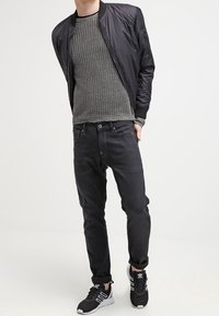 Giubbotto bomber nero su un maglione grigio lavorato a maglia, con jeans scuri e sneakers nere con strisce bianche. Design semplice e moderno.