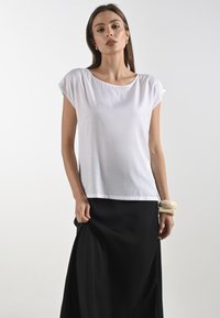 Top bianco a maniche corte con scollo rotondo, realizzato in tessuto morbido, abbinato a una gonna nera fluente. Bracciale largo di colore chiaro al polso.