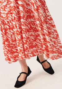 Maxikleid mit rot-weißen Muster und ausgestellt geschnittenem Saum; schwarze Mary Jane Ballerinas mit Riemen und niedrigem Absatz. Glatte Stoffoberfläche.