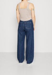 Džínové kalhoty s širokými nohavicemi v tmavě modrém odstínu, s dvěma zadními kapsami, prošitými detaily a vysokým pasem, v kombinaci s béžovým topem bez rukávů.