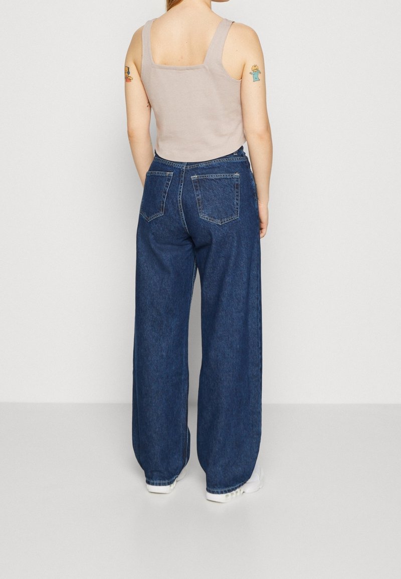 Džínové kalhoty s širokými nohavicemi v tmavě modrém odstínu, s dvěma zadními kapsami, prošitými detaily a vysokým pasem, v kombinaci s béžovým topem bez rukávů.