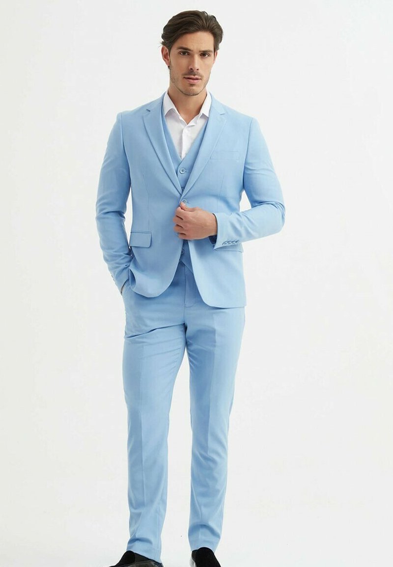 Un costume bleu clair à la coupe ajustée, comprenant un blazer à deux boutons, des revers à cran et un pantalon à plis plats. Chemise blanche en dessous, chaussures noires.