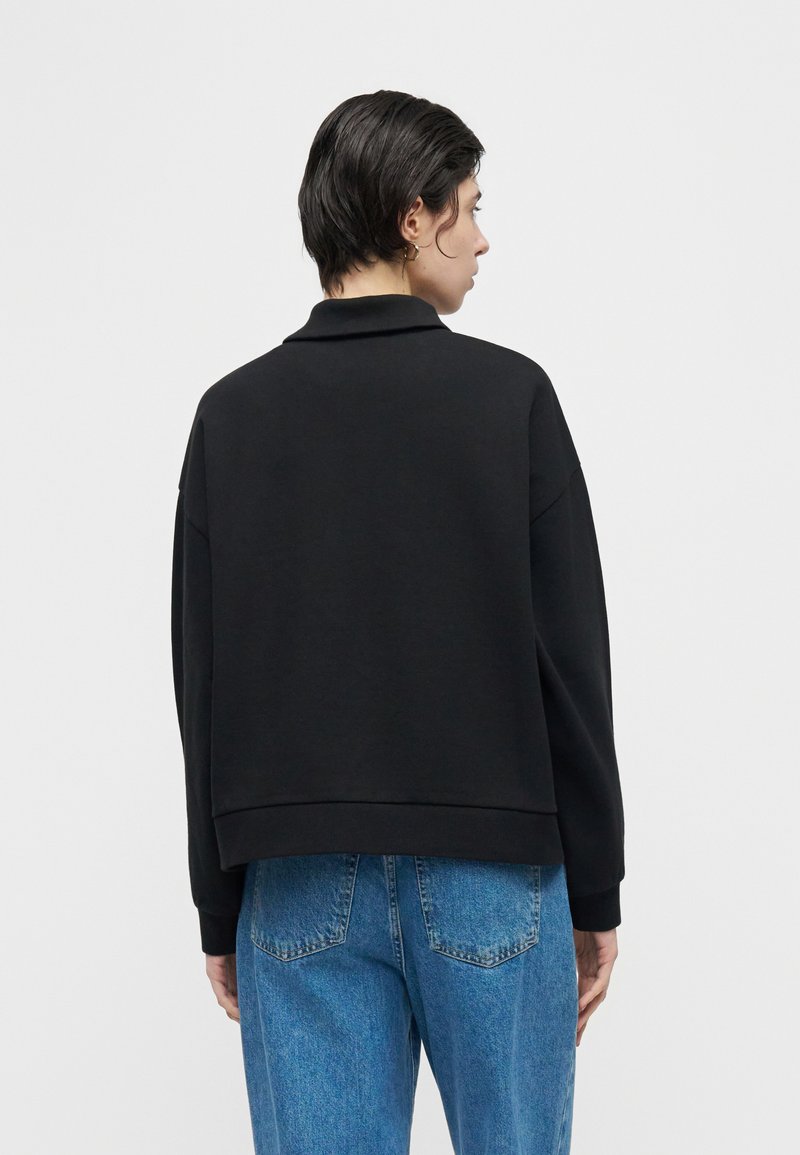 Personne aux cheveux courts et foncés, vêtue d'un haut noir à manches longues et d'un jean bleu, vue de dos sur un fond blanc uni.