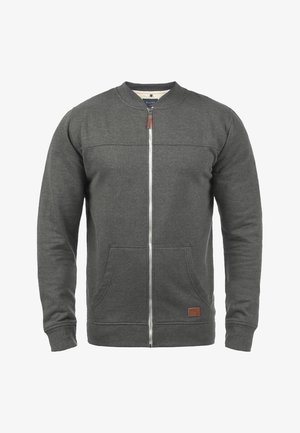 Harmaa vetoketjullinen huppari, jossa on pyöreä kaulus, kaksi etutaskua, sileä pinta ja nahkainen korostelappu helman lähellä.