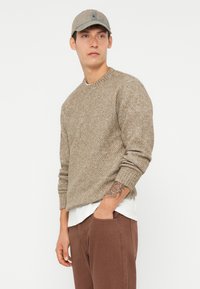Beige knit sweater with a textured pattern, round neckline, ribbed cuffs, worn over a white shirt. Paired with brown relaxed-fit trousers.