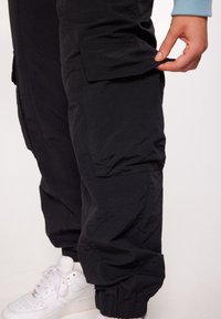 Zwarte cargo broek met een verfrommelde textuur, voorzien van een zijzak en elastische boorden, gecombineerd met witte sneakers.