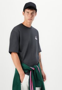 Grå bomulds t-shirt med korte ærmer og rund halsudskæring, med et lille lyserødt logo på brystet. Grøn sweatshirt bundet om taljen.