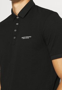 Svart pikétröja i bomull med texturerad tyg, med en tvåknapps knäppning och kontrasterande krage. Litet logotyp på bröstet.