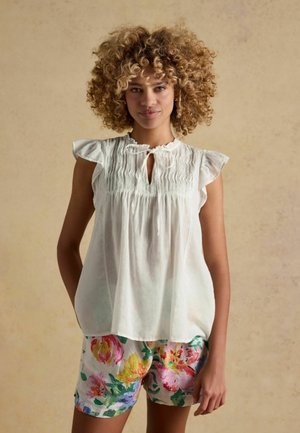Kobieta o kręconych włosach, ubrana w białą, falbaniaste, bezrękawnikową bluzkę i kolorowe, kwiatowe szorty, stojąca na beżowym tle.