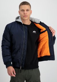 Marinfärgad bomberjacka med grå huva, orange foder och ribbade muddarna. Dragkedjestängning och logotyp på bröstet. Bärs över en svart sweatshirt.